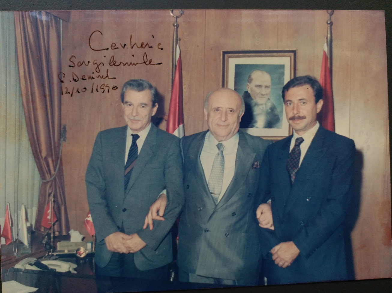 Süleyman Demirel, arkada Atatürk portresi ve bayrakların bulunduğu ahşap kaplı bir makam odasında iki kişiyle kol kola poz veriyor. Fotoğrafın sol üst köşesinde Demirel'in el yazısıyla “Cevher'e sevgilerimle, S. Demirel, 12/10/1990” ithafı var.